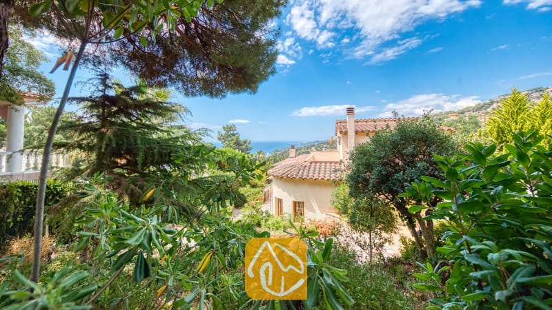 Vakantievilla's Costa Brava Spanje - Villa Cleo - Buitenaanzicht van Villa Cleo in Spanje, omlijst door weelderig groen tuingroen met traditionele terracotta dakpannen en de zee zichtbaar op de achtergrond onder een helderblauwe hemel.