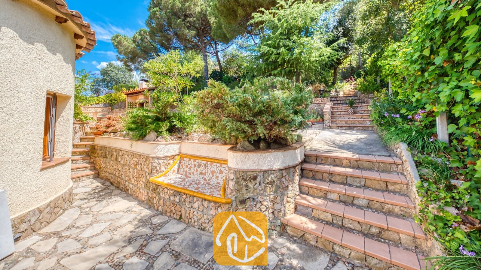 Vakantievilla's Costa Brava Spanje - Villa Cleo - Stenen terras bij Villa Cleo met stenen trappen, keermuren met een mozaïektegelbank en weelderig omliggend groen onder een blauwe hemel
