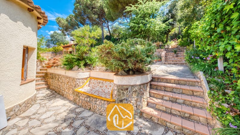 Vakantievilla's Costa Brava Spanje - Villa Cleo - Stenen terras bij Villa Cleo met stenen trappen, keermuren met een mozaïektegelbank en weelderig omliggend groen onder een blauwe hemel