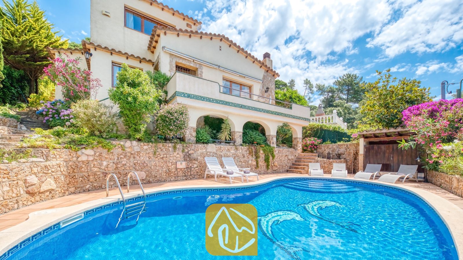 Vakantievilla's Costa Brava Spanje - Villa Cleo - Buitenaanzicht van Villa Cleo met een blauw zwembad voorzien van dolfijnmozaïek, witte ligstoelen, stenen keermuren en een meertraps crème kleurige villa onder een bewolkte hemel.