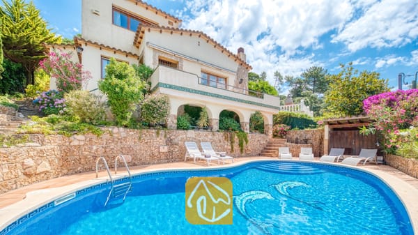 Vakantievilla's Costa Brava Spanje - Villa Cleo - Buitenaanzicht van Villa Cleo met een blauw zwembad voorzien van dolfijnmozaïek, witte ligstoelen, stenen keermuren en een meertraps crème kleurige villa onder een bewolkte hemel.