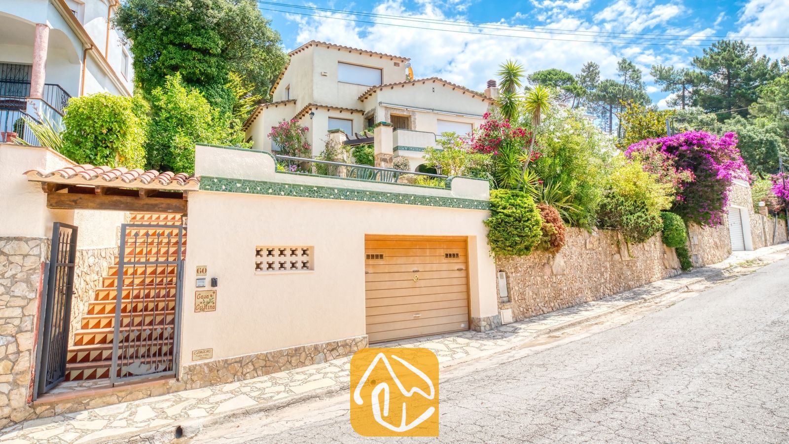 Vakantievilla's Costa Brava Spanje - Villa Cleo - Buitenaanzicht van Villa Cleo met een beige muur, een houten garagedeur, een trap met motieftaljels en een metalen hek, omringd door groen onder een blauwe lucht.