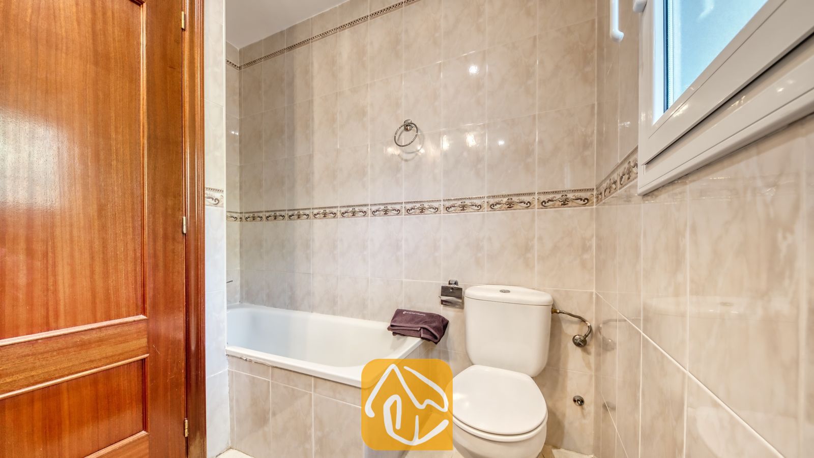Vakantievilla's Costa Brava Spanje - Villa Chanel - Badkamer voorzien van een wit porseleinen ligbad en toilet, neutrale beige tegels met een decoratieve rand en een gepolijste houten deur.