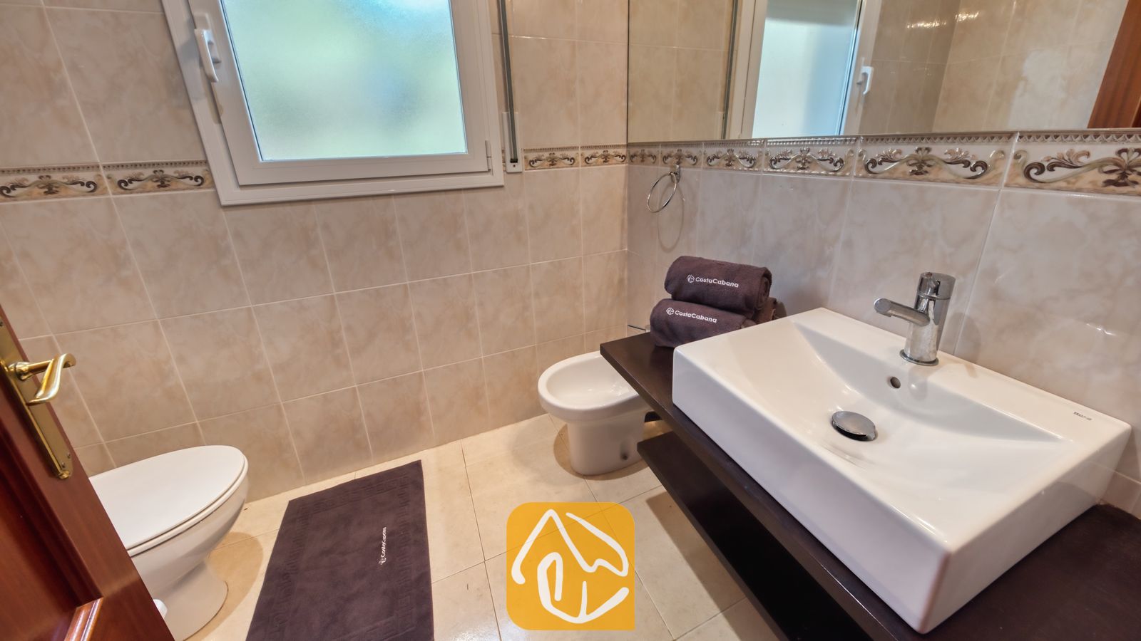 Vakantievilla's Costa Brava Spanje - Villa Chanel - Een witte waskom op een donker wastafelmeubel in een badkamer van Villa Chanel, voorzien van beige wandtegels, een bidet en een matglazen raam.