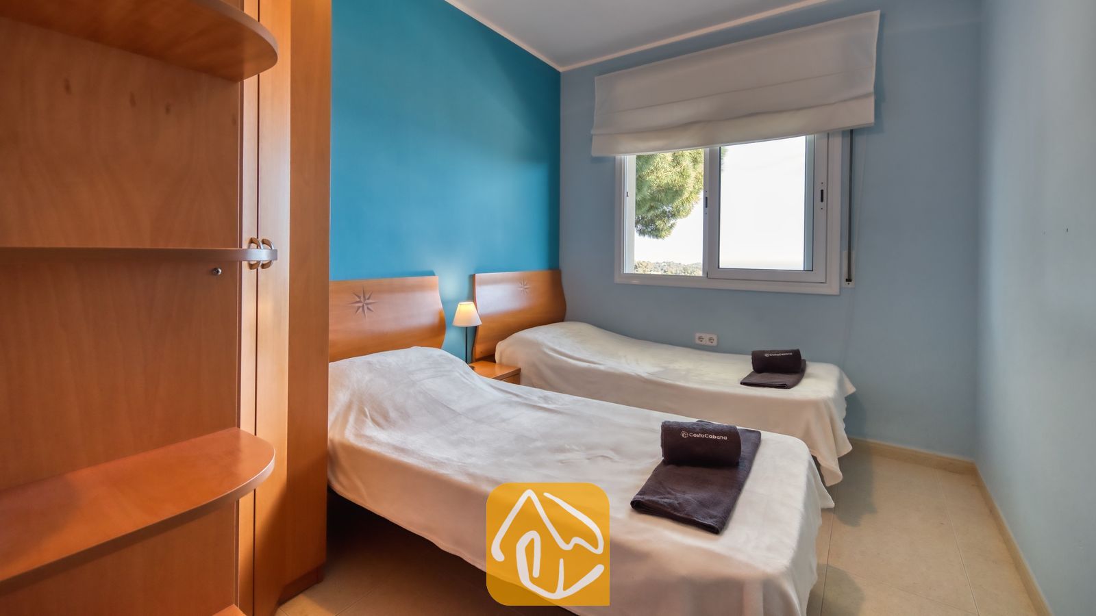 Vakantievilla's Costa Brava Spanje - Villa Chanel - Tweepersoonskamer in Villa Chanel, voorzien van twee eenpersoonsbedden met wit bedlinnen, houten hoofdborden en een blauwe accentmuur naast een raam met witte jaloezieën.
