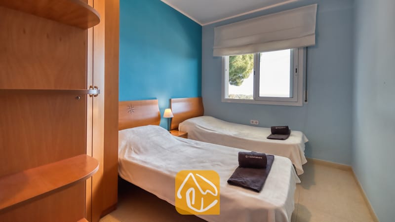 Villas de vacances Costa Brava Espagne - Villa Chanel - Chambre double à la Villa Chanel, équipée de deux lits simples avec linge de maison blanc, têtes de lit en bois et un mur d'accent bleu situé près d'une fenêtre habillée d'un store blanc.