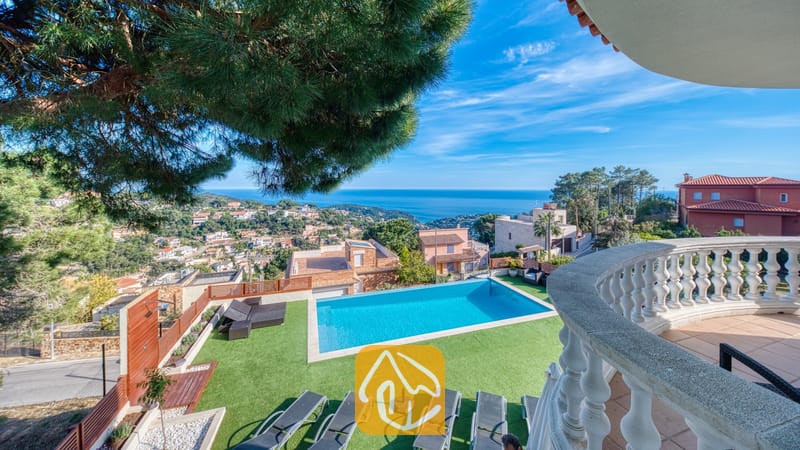 Vakantievilla's Costa Brava Spanje - Villa Chanel - Privézwembad bij Villa Chanel met grijze ligbedden op een groen gazon, gezien vanaf een stenen balkon met uitzicht op de zee en de heuvels langs de kust.