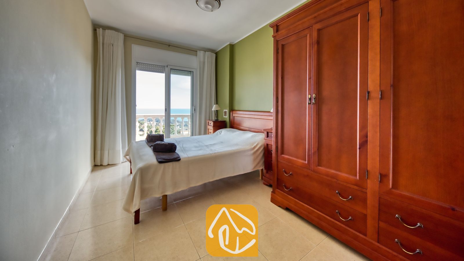 Vakantievilla's Costa Brava Spanje - Villa Chanel - Slaapkamer met een grote houten kledingkast, een groene accentmuur en een bed met wit bedlinnen. Glazen deuren bieden toegang tot een balkon met uitzicht op zee bij Villa Chanel.