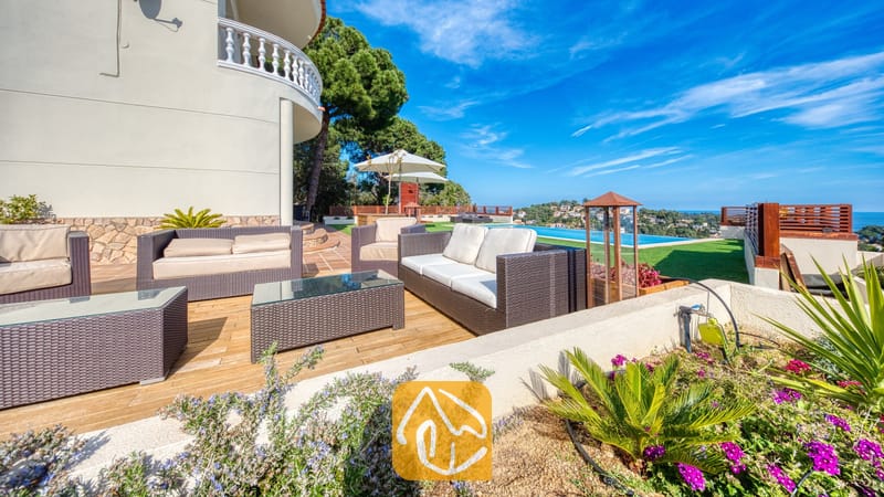 Vakantievilla's Costa Brava Spanje - Villa Chanel - Terraslounge bij Villa Chanel, voorzien van donker vlechtwerkmeubilair met crèmekleurige kussens, glazen tafels en een infinity pool met uitzicht op zee.