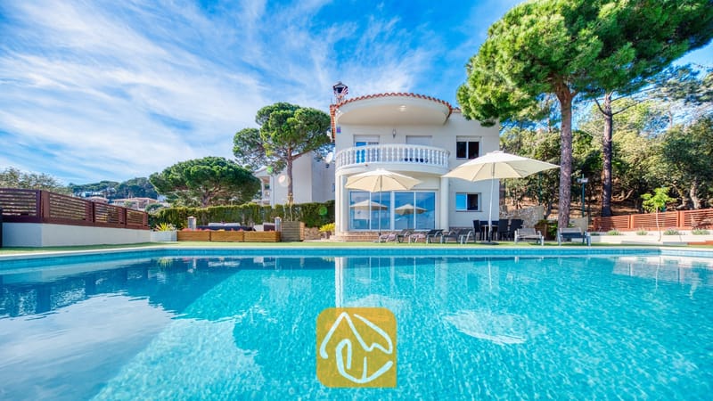 Vakantievilla's Costa Brava Spanje - Villa Chanel - De buitenkant van Villa Chanel met een turquoise zwembad, witte parasols op het terras en een gebogen stenen balkon, omringd door weelderige groene dennenbomen.
