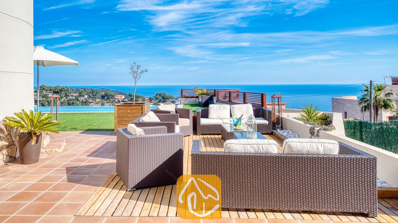 Vakantievilla's Costa Brava Spanje - Villa Chanel - Terras van Villa Chanel met bruin vlechtwerk loungeset, witte kussens en een houten vlonder met uitzicht op zee en een infinity pool.