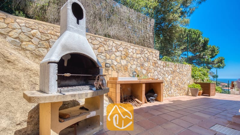 Vakantievilla's Costa Brava Spanje - Villa Chanel - Buitenbarbecue van natuursteen en een spoelbak op een betegeld terras bij Villa Chanel, gekenmerkt door een stenen muur en een prachtig uitzicht op de verre zee.