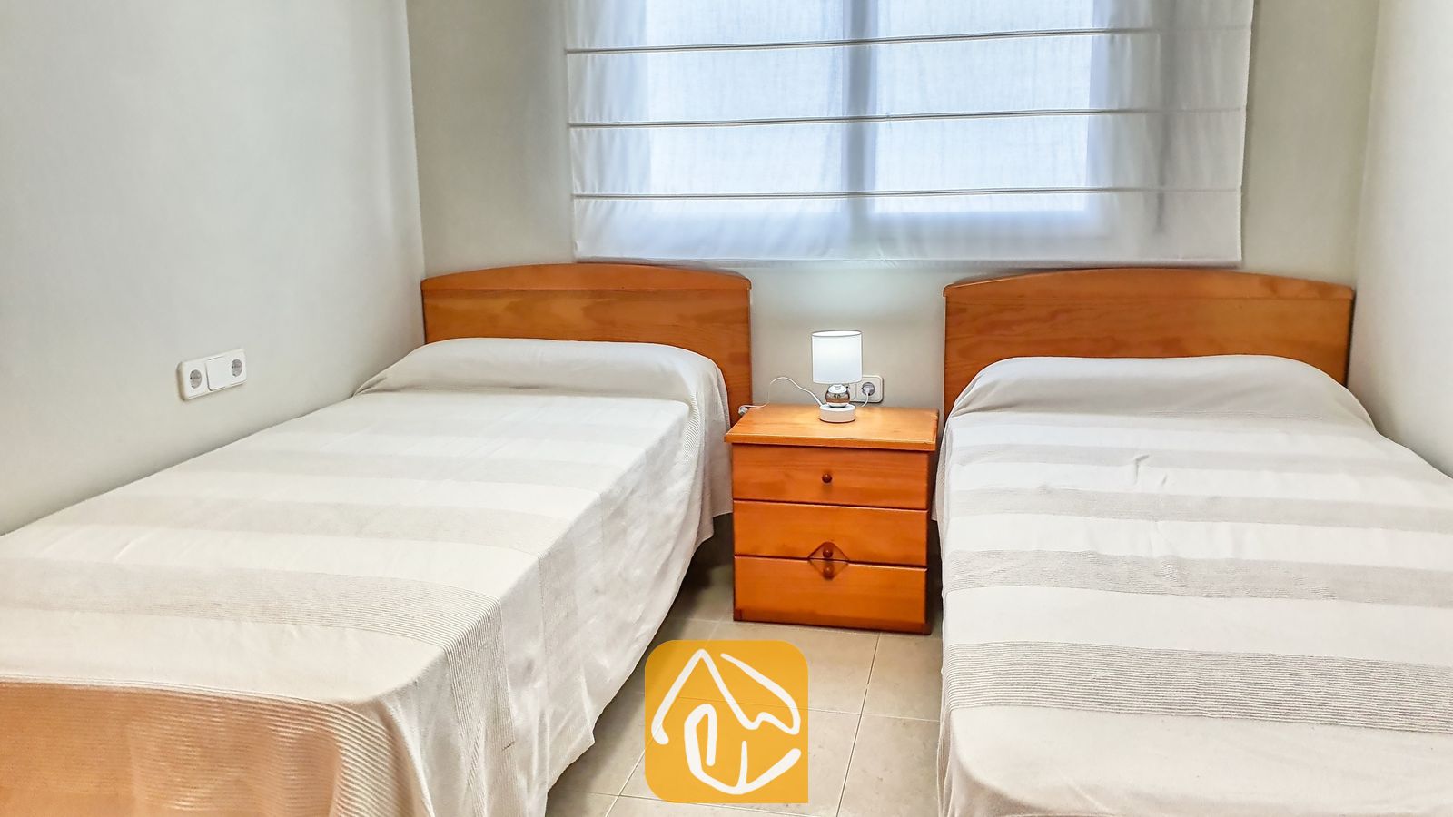 Vakantievilla's Costa Brava Spanje - Villa Chanel - Slaapkamer met twee eenpersoonsbedden voorzien van houten hoofdborden, wit gestreept beddengoed en een centraal nachtkastje met een klein lampje in Villa Chanel.