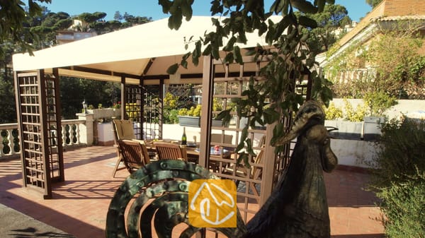 Vakantievilla's Costa Brava Spanje - Villa Noa - Houten pergola met een stoffen doek op een zonnig terras bij Villa Noa, voorzien van een eettafel en stoelen in de schaduw.