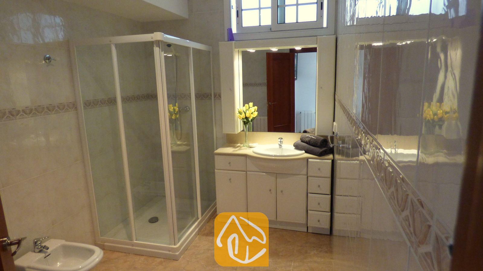 Vakantievilla's Costa Brava Spanje - Villa Noa - Badkamer met een hoekdouche, een wit badmeubel met wastafel, een verlichte spiegel en gele bloemen in een vaas.