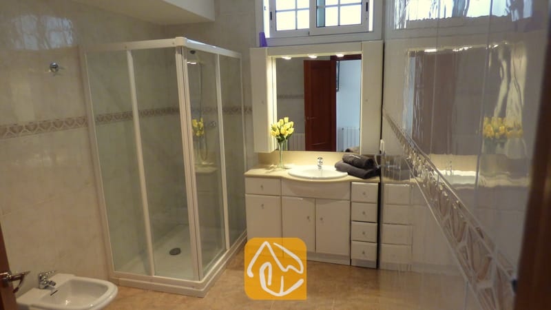 Holiday villas Costa Brava Spain - Villa Noa - Bathroom with a corner shower enclosure, white vanity cabinet with a sink, lighted mirror, and yellow flowers in a vase.