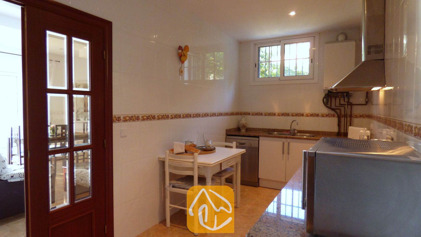 Vakantievilla's Costa Brava Spanje - Villa Noa - Keuken met witte betegelde muren, een kleine witte eettafel met twee stoelen en een donkere houten paneeldeur.