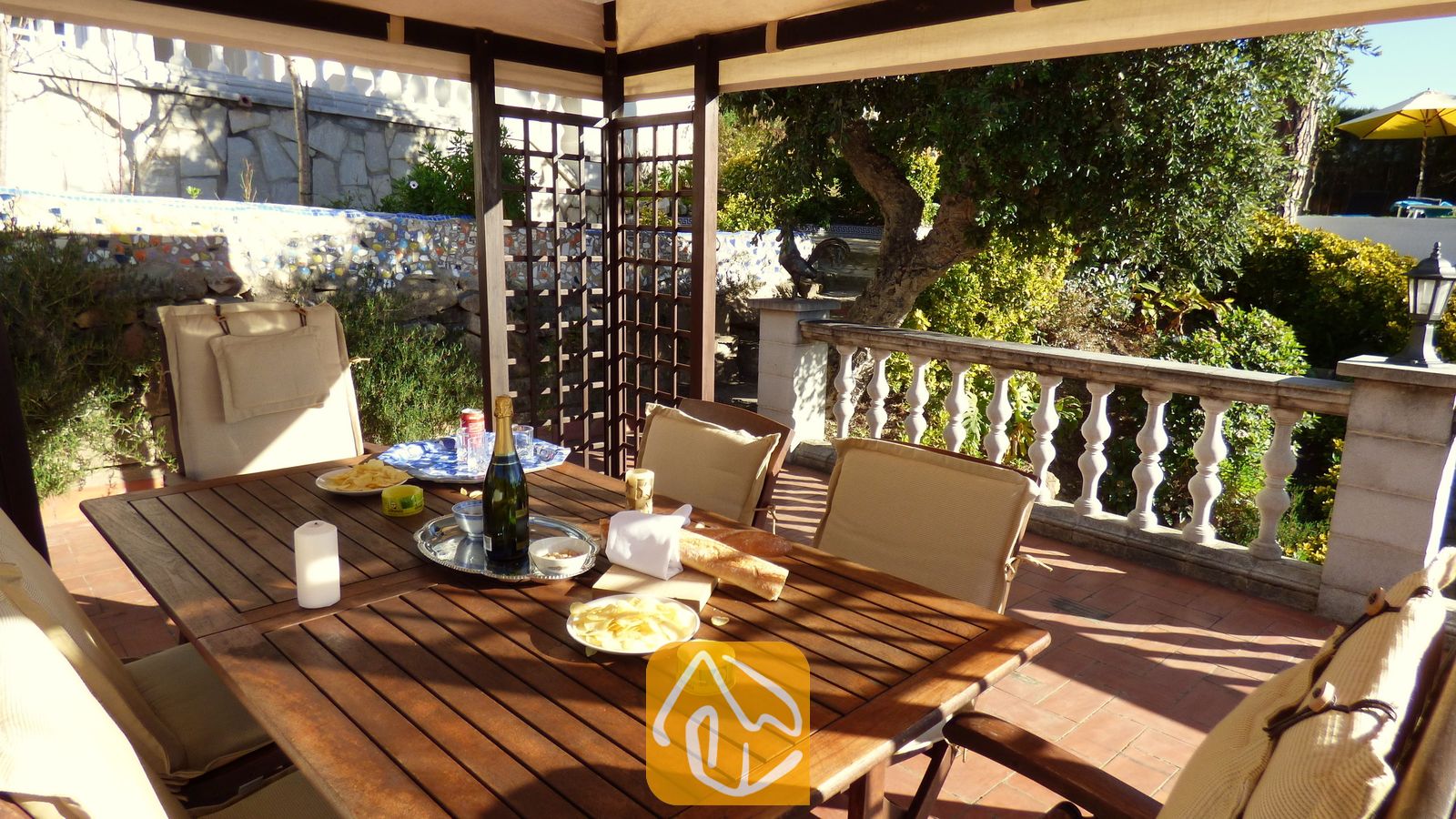 Vakantievilla's Costa Brava Spanje - Villa Noa - Overdekt houten eetterras bij Villa Noa met een tafel, gedekt met wijn, snacks en kaarsen, en uitzicht op een tuin met een stenen balustrade.