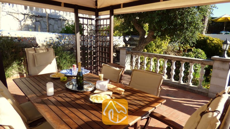 Vakantievilla's Costa Brava Spanje - Villa Noa - Overdekt houten eetterras bij Villa Noa met een tafel, gedekt met wijn, snacks en kaarsen, en uitzicht op een tuin met een stenen balustrade.