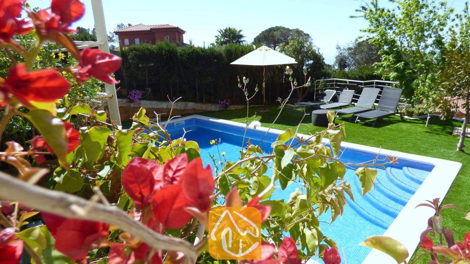 Vakantievilla's Costa Brava Spanje - Villa Noa - Het privézwembad bij Villa Noa, omlijst door rode bougainvillea, met ligstoelen en een witte parasol op het gazon op de achtergrond.