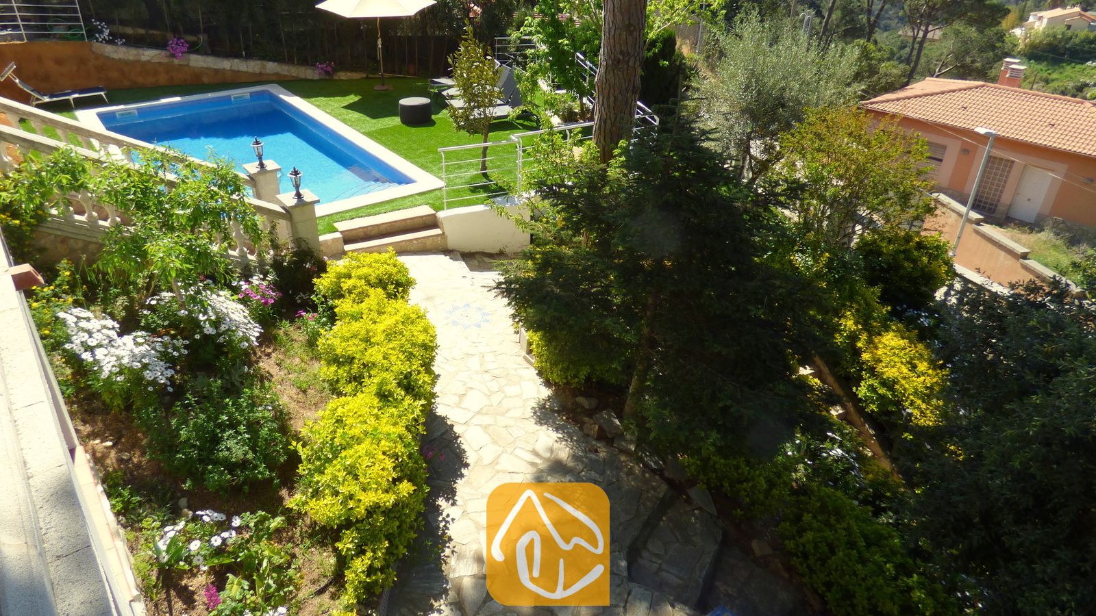 Vakantievilla's Costa Brava Spanje - Villa Noa - Hoge hoekperspectief van Villa Noa met een stenen tuinpad, weelderige groene struiken, bloeiende planten en een rechthoekig zwembad omringd door kunstgras.