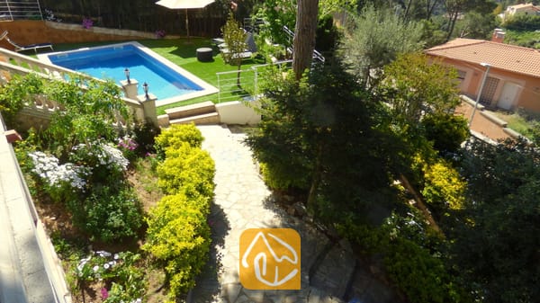 Vakantievilla's Costa Brava Spanje - Villa Noa - Hoge hoekperspectief van Villa Noa met een stenen tuinpad, weelderige groene struiken, bloeiende planten en een rechthoekig zwembad omringd door kunstgras.
