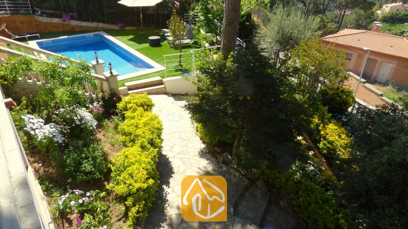 Vakantievilla's Costa Brava Spanje - Villa Noa - Hoge hoekperspectief van Villa Noa met een stenen tuinpad, weelderige groene struiken, bloeiende planten en een rechthoekig zwembad omringd door kunstgras.