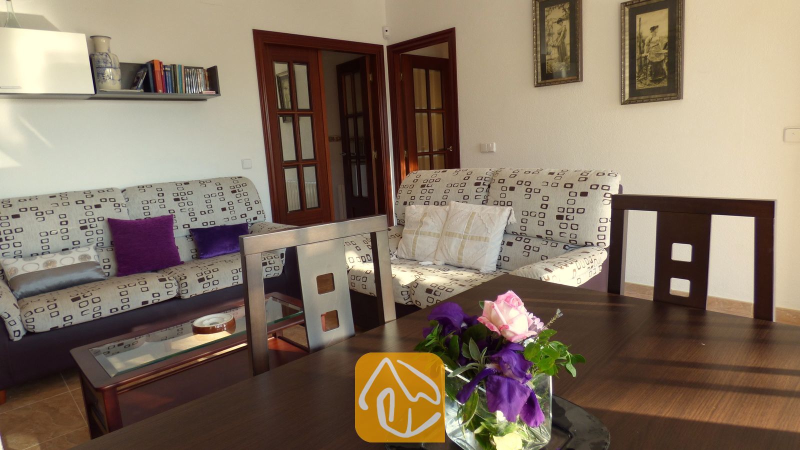 Vakantievilla's Costa Brava Spanje - Villa Noa - Woonkamer met twee patroonbanken, houten deuren, ingelijste muurkunst en op de voorgrond een donkerhouten eettafel.