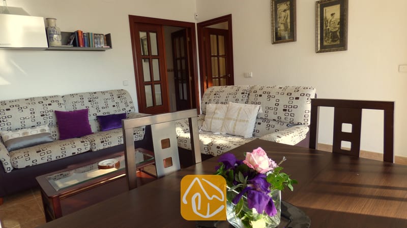 Vakantievilla's Costa Brava Spanje - Villa Noa - Woonkamer met twee patroonbanken, houten deuren, ingelijste muurkunst en op de voorgrond een donkerhouten eettafel.