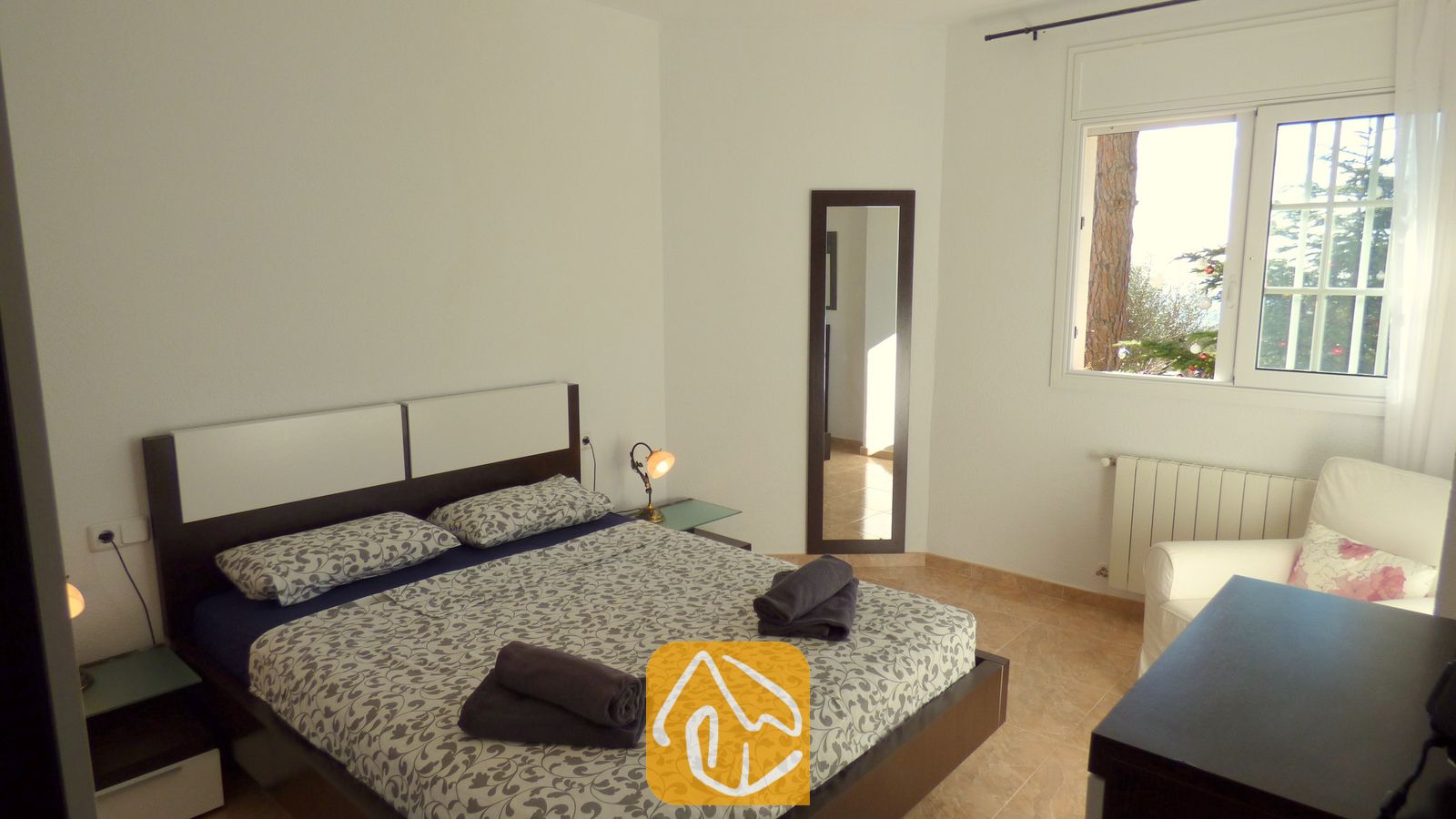 Vakantievilla's Costa Brava Spanje - Villa Noa - Slaapkamer met een tweepersoonsbed voorzien van grijs beddengoed met motief, twee opgerolde donkere handdoeken, een nachtkastjelamp, een passpiegel en een raam met uitzicht op bomen.