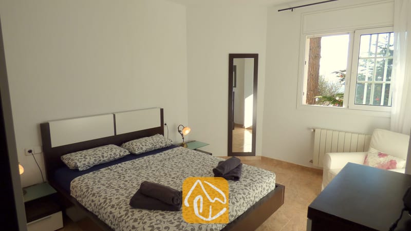 Holiday villas Costa Brava Spain - Villa Noa - Bedroom with a double bed featuring patterned grey bedding, two rolled dark towels, a bedside lamp, a full-length mirror, and a window with tree views.