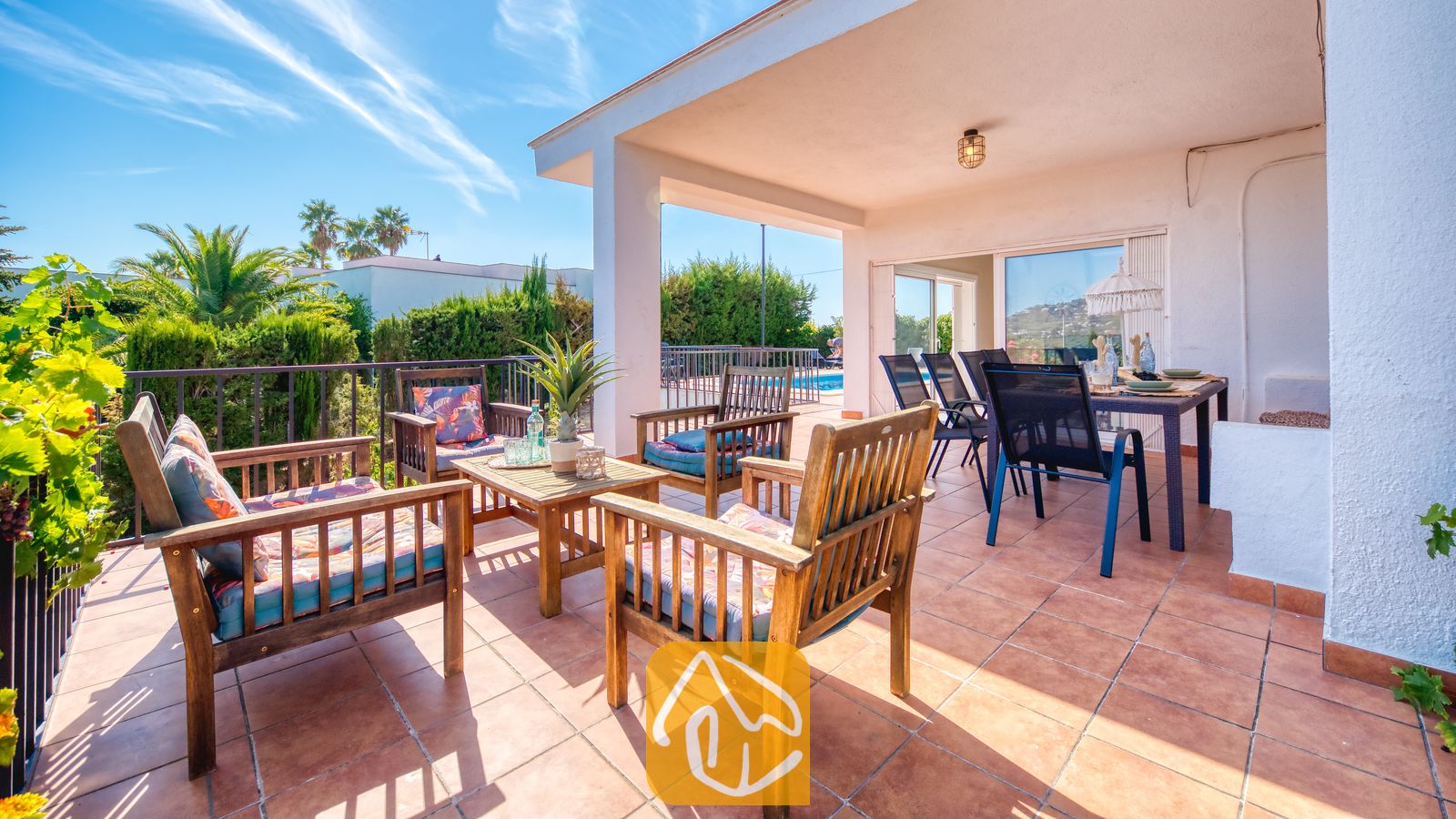 Holiday villas Costa Brava Spain - Villa Yara - Covered terrace at Villa Yara featuring wooden armchairs with patterned cushions, a coffee table, and an outdoor dining area overlooking palm trees.