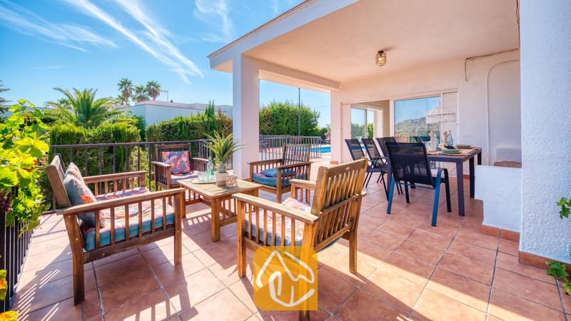 Holiday villas Costa Brava Spain - Villa Yara - Covered terrace at Villa Yara featuring wooden armchairs with patterned cushions, a coffee table, and an outdoor dining area overlooking palm trees.