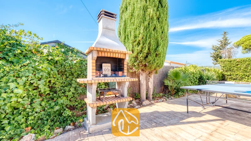 Holiday villas Costa Brava Spain - Villa Yara - Outdoor stone barbecue grill with a brick-trimmed hearth standing on a tiled patio at Villa Yara, with green foliage and a blue ping pong table nearby.
