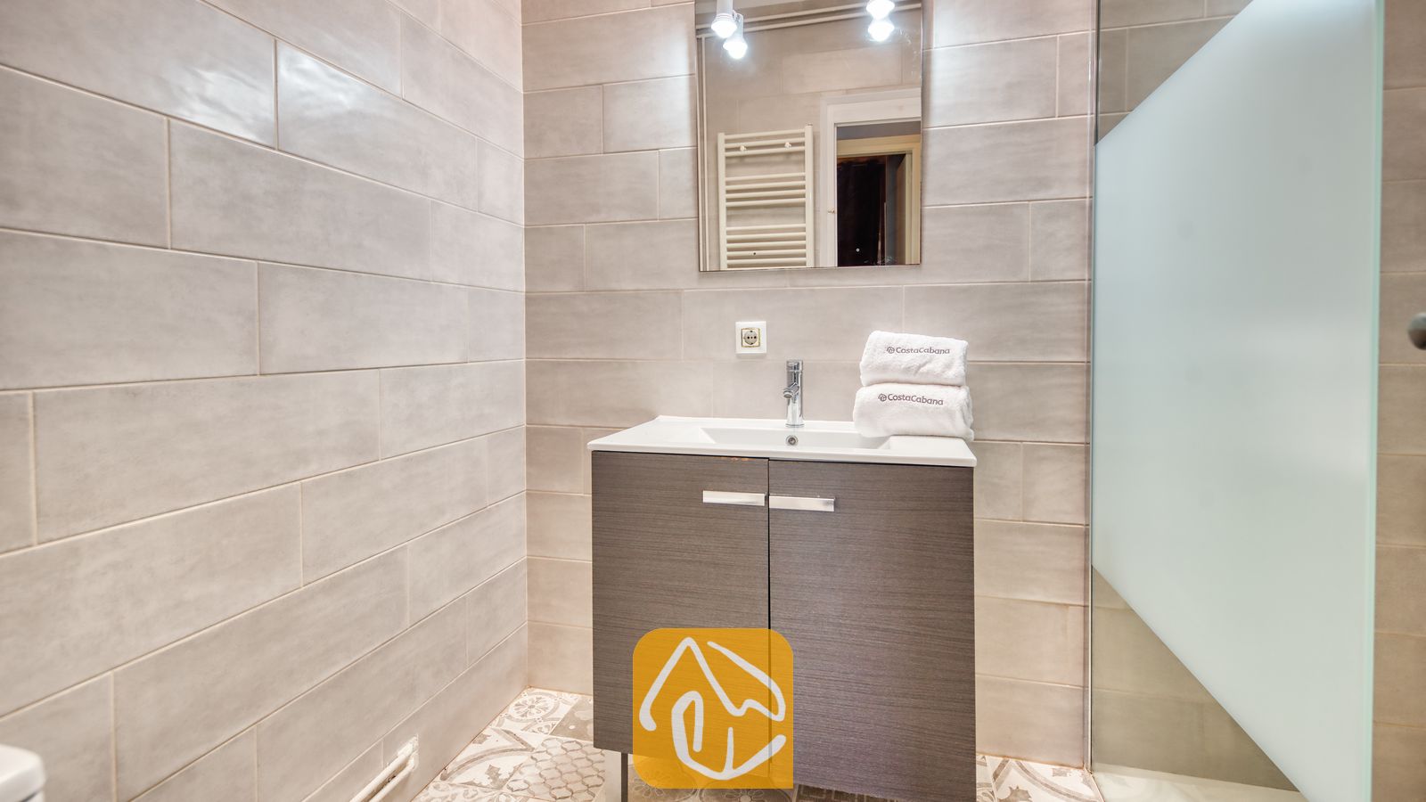 Holiday villas Costa Brava Spain - Villa Yara - Modern bathroom vanity with a white basin and folded towels underneath a rectangular mirror against grey tiled walls.