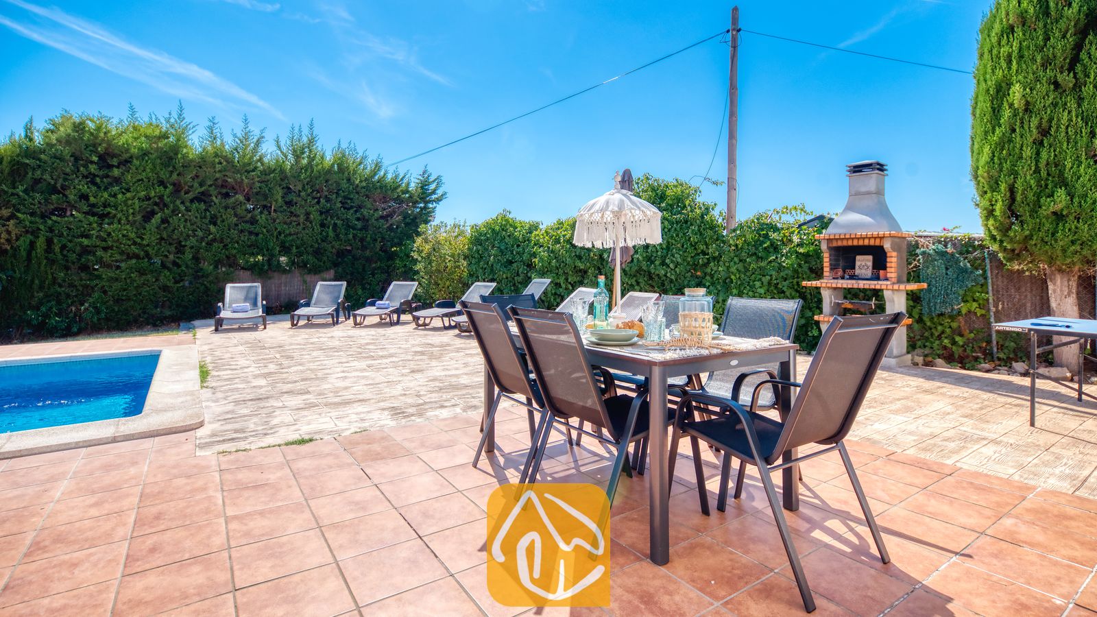 Holiday villas Costa Brava Spain - Villa Yara - Outdoor dining table with dark metal chairs, small decorative parasol, and glassware on a tiled terrace at Villa Yara, alongside a swimming pool and brick barbecue grill.