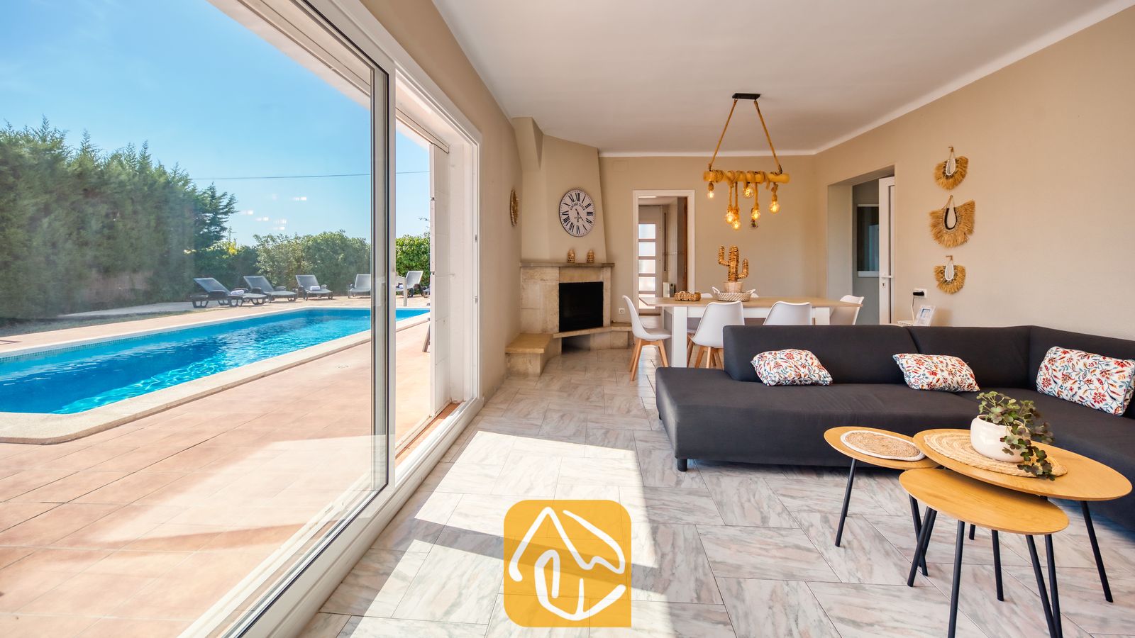 Holiday villas Costa Brava Spain - Villa Yara - Villa Yara living room with sliding glass doors opening to a swimming pool, dark grey sofa, light wood nesting coffee tables, and a corner fireplace.