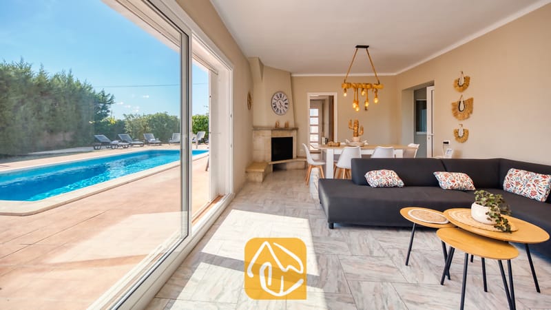 Holiday villas Costa Brava Spain - Villa Yara - Villa Yara living room with sliding glass doors opening to a swimming pool, dark grey sofa, light wood nesting coffee tables, and a corner fireplace.