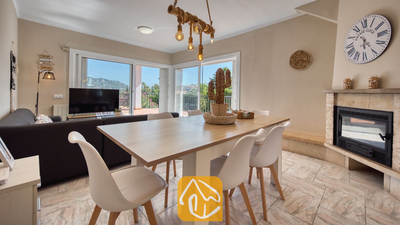 Holiday villas Costa Brava Spain - Villa Yara - Wooden dining table with white chairs inside Villa Yara, featuring a fireplace, large wall clock, and sliding glass doors opening to a terrace.