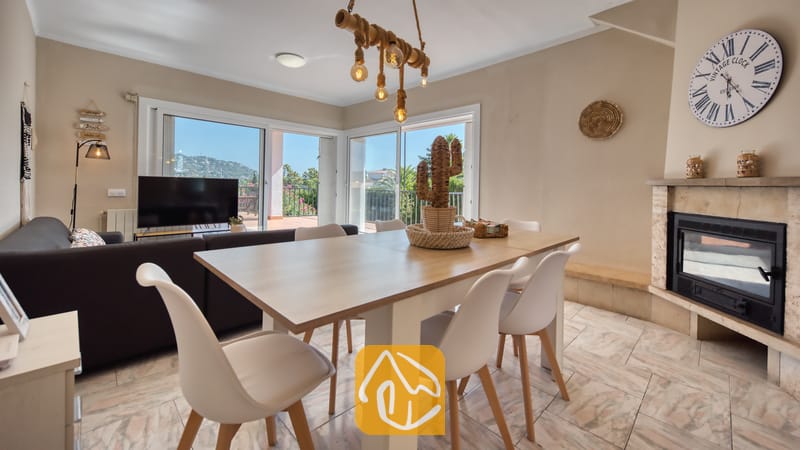 Holiday villas Costa Brava Spain - Villa Yara - Wooden dining table with white chairs inside Villa Yara, featuring a fireplace, large wall clock, and sliding glass doors opening to a terrace.