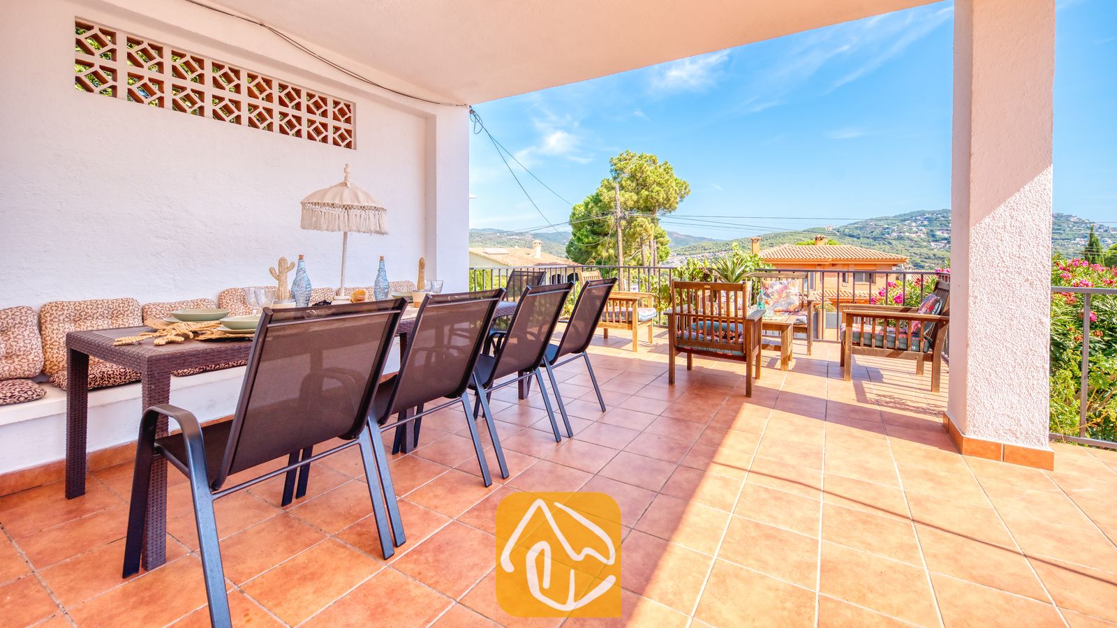 Holiday villas Costa Brava Spain - Villa Yara - Covered terrace at Villa Yara featuring a dining table with mesh chairs, tiled flooring, and a view of surrounding hills and trees under a blue sky.