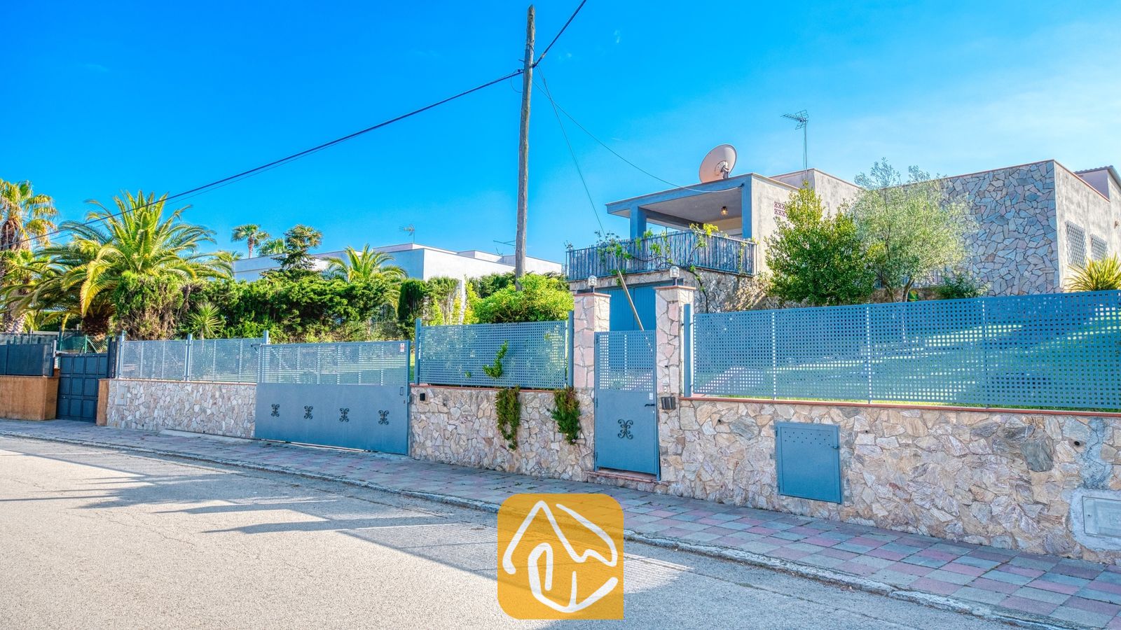 Vakantievilla's Costa Brava Spanje - Villa Yara - Buitengevel van Villa Yara met een stenen muur, blauw metalen hek en de voorste poort langs de straat onder een helderblauwe hemel.