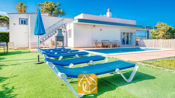 Vakantievilla's Costa Brava Spanje - Villa Yara - Buitenaanzicht van Villa Yara met blauwe ligstoelen op kunstgras naast een rechthoekig zwembad en een wit gebouw van twee verdiepingen.