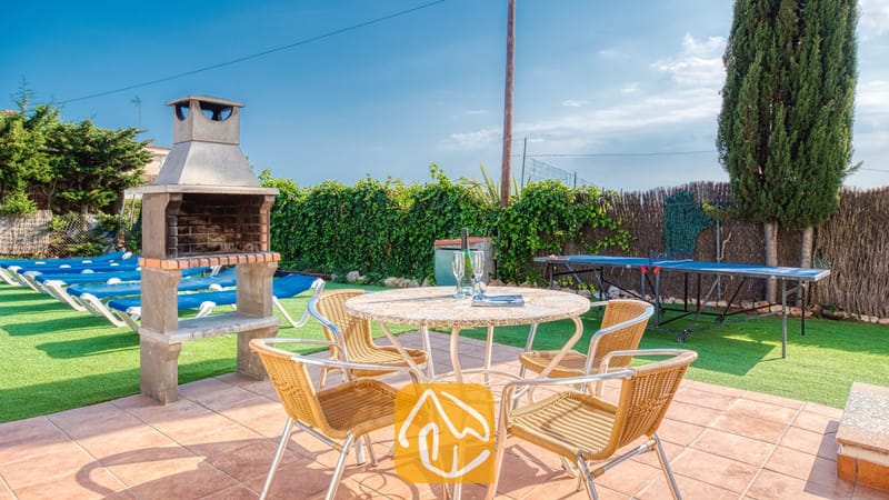 Vakantievilla's Costa Brava Spanje - Villa Yara - Eethoek in de buitenlucht bij Villa Yara, voorzien van een stenen barbecue, een ronde stenen tafel met vier geweven stoelen en een tafeltennistafel op het grasveld.