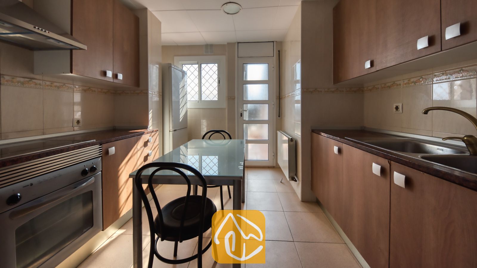 Vakantievilla's Costa Brava Spanje - Villa Yara - Smalle keuken met houten kasten, een glazen eettafel met twee stoelen, een roestvrijstalen oven en een tegelvloer.
