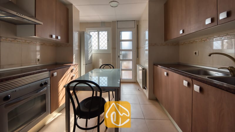 Vakantievilla's Costa Brava Spanje - Villa Yara - Smalle keuken met houten kasten, een glazen eettafel met twee stoelen, een roestvrijstalen oven en een tegelvloer.