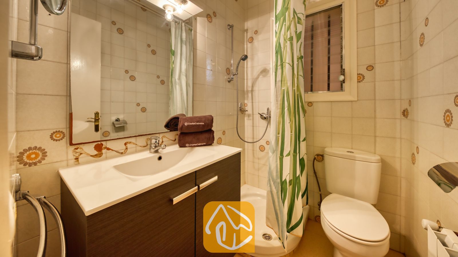 Vakantievilla's Costa Brava Spanje - Villa Yara - Badkamer met een wastafelmeubel van donker hout met een witte wasbak, een spiegel aan de muur, een hoekdouche met een groen geprint gordijn en een wit toilet te midden van muren met gedessineerde tegels.