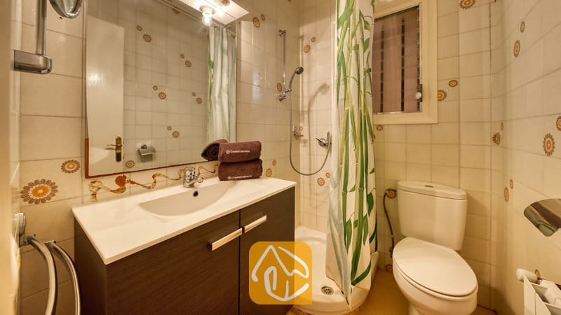 Holiday villas Costa Brava Spain - Villa Yara - Bathroom featuring a dark wood vanity with a white basin, a wall mirror, a corner shower with a green patterned curtain, and a white toilet against patterned tiled walls.