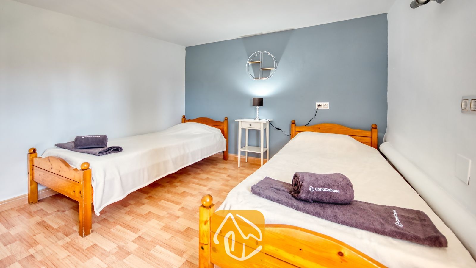 Vakantievilla's Costa Brava Spanje - Villa Yara - Slaapkamer met twee houten eenpersoonsbedden, wit beddengoed, opgevouwen grijze handdoeken, een klein wit nachtkastje en een ronde wandplank tegen een blauwe accentmuur.