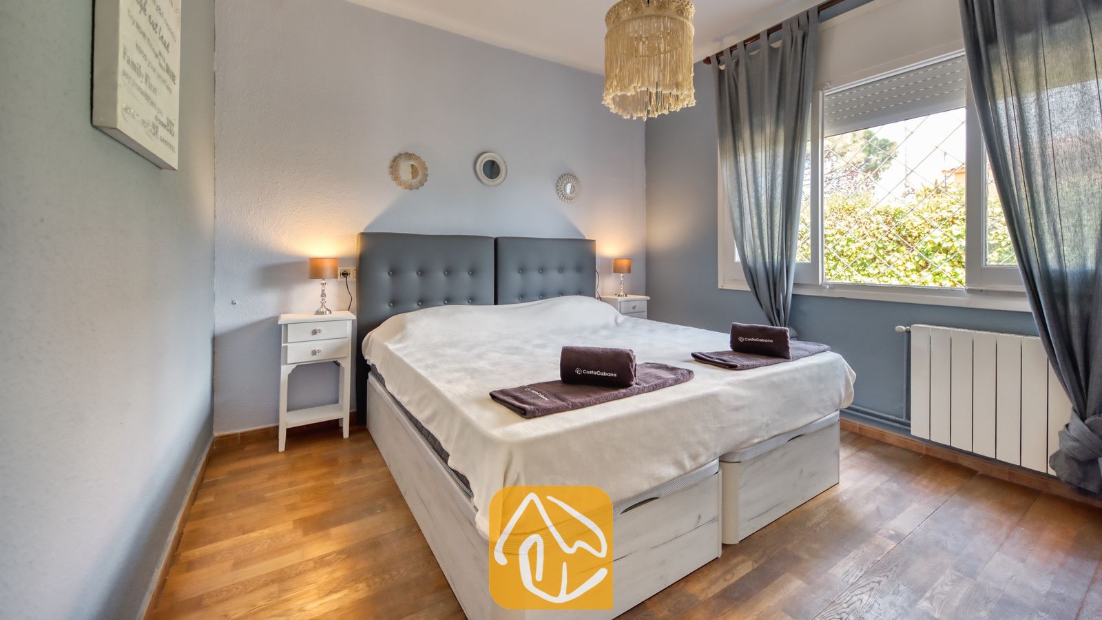 Vakantievilla's Costa Brava Spanje - Villa Yara - Slaapkamer met een donkergrijs gestoffeerd hoofdbord, witte nachtkastjes met lampen, en opgerolde bruine handdoeken op het bed onder een hanglamp met franjes.